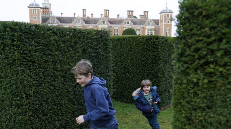Two children dressed in blue winter wear playing amongst the clipped hedges in the grounds with Blickling Hall in the backgroound